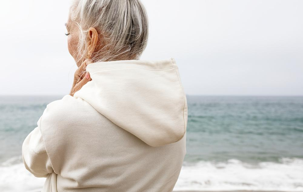 Reife Frau mit grauen Haaren im weissen Hoodie schaut zum Meer, von hinten fotografiert