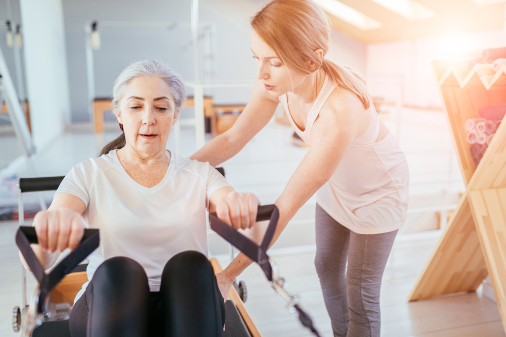 Trainerin leitet eine reife Dame bei einer Uebung auf dem Pilates Reformer
