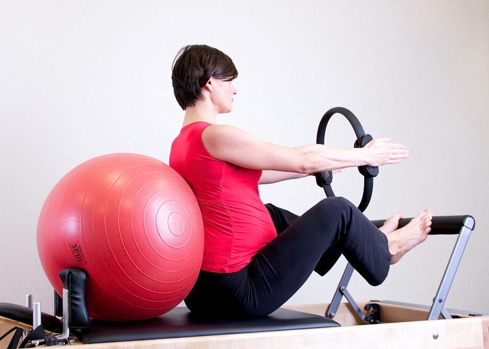 Schwangere Frau mit roten T-shirt auf Pilates Reformer sitzend gegen einem Fitness Ball und mit dem Pilates Ring zwischen ihrer Haende